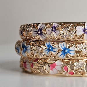 May include: Three gold-toned bangles stacked on top of each other. Each bangle features a floral design with white and colored enamel flowers, accented with small clear crystals. The colors of the flowers are purple, blue, and pink.