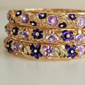 May include: Three gold-toned bangles stacked, adorned with purple and lavender floral designs. Each flower has a central rhinestone. The bangles have an openwork design with green leaves.