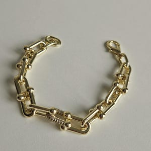 May include: A gold-toned chain bracelet featuring elongated rectangular links connected by small spheres. One link is encrusted with small clear stones. The bracelet includes a lobster claw clasp and a small circular ring at the opposite end.