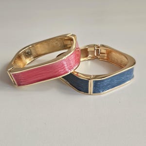 May include: Two gold-toned metal hinged bracelets. One bracelet has a pink enamel inlay, and the other has a blue enamel inlay. The bracelets have a square shape with rounded corners. The bracelets are displayed on a white surface.