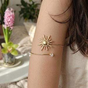 Puede incluir: Brazalete dorado con diseño de sol y elemento circular central. El brazalete tiene un estilo delicado y minimalista, con un pequeño acento de piedra preciosa transparente. La joya se lleva en la parte superior del brazo, añadiendo un toque de elegancia.