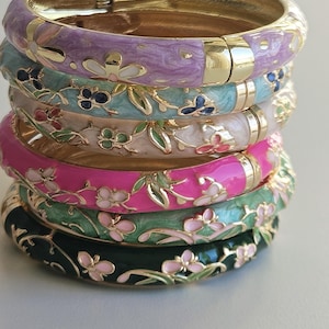 Può includere: Pila di sette braccialetti smaltati colorati. Ogni braccialetto è di un colore diverso, tra cui viola, verde acqua, rosa, verde e nero. I braccialetti sono decorati con disegni floreali dorati e hanno una chiusura dorata.