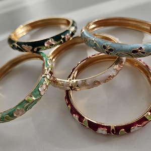 Peut inclure: Cinq bracelets dorés ornés de motifs floraux en émail de différentes couleurs : vert foncé, bleu clair, vert menthe, blanc et bordeaux. Les bracelets sont disposés sur une surface blanche, mettant en valeur leurs détails.