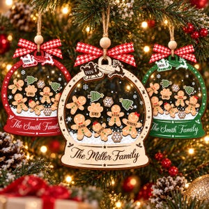 Könnte beinhalten: Drei kugelförmige Weihnachtsschmuckstücke mit Holzrahmen in Rot, Naturholz und Grün. Jedes Ornament zeigt Lebkuchenfiguren, Pfotenabdrücke, Bäume, die Jahreszahl 2026 sowie personalisierte Familiennamen auf der Vorderseite.