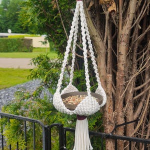 Op de afbeelding: Een handgemaakte macrame plantenhouder in wit, met een donkerbruine kom gevuld met zaden. De hanger heeft een bordeauxrode accent en een lange kwast. De plantenhouder hangt aan een metalen haak, tegen een achtergrond van groen gebladerte.