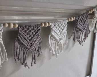 May include: A macrame garland featuring alternating triangular pennants in cream and grey cord. Each pennant displays a knotted diamond pattern with long fringe at the bottom. The pieces are strung together on a cord interspersed with round wooden beads.