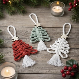 Può includere: Tre ornamenti natalizi in macramè fatti a mano in rosso, verde e bianco. Ogni albero ha una stella di legno e un nappa bianca. Gli ornamenti sono esposti su una superficie di legno con verde festivo e candele.