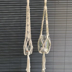 Può includere: Due supporti per piante in macramè color bianco sporco appesi a dei ganci. Il supporto sinistro è sottile con un piccolo portacandele in vetro. Quello destro è più largo, con accenti verde salvia e un barattolo di vetro, entrambi con nappe.