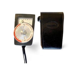 May include: A vintage Sekonic light meter with a black body and a circular dial featuring ASA and DIN markings. The meter is accompanied by a black leather case with the Sekonic logo embossed on it. A black strap is attached to the meter.