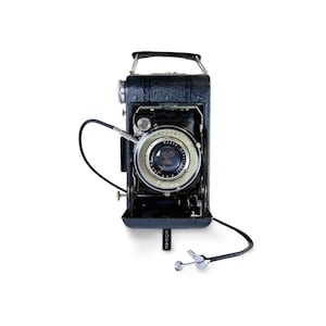 May include: A vintage black Kodak camera with a silver lens and a black handle. The camera has a bellows design and a cable release attached. The Kodak logo is visible on the bottom of the camera.