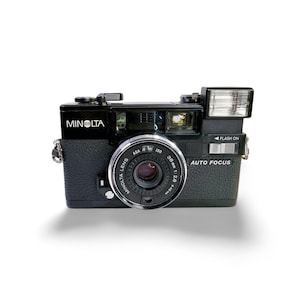 Puede incluir: Cámara de película Minolta negra de 35 mm con lente plateada y flash incorporado. La lente está marcada con "Minolta Lens" y "38mm 1:2.8". La cámara tiene las etiquetas "Auto Focus" y "Flash On".
