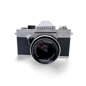 Puede incluir: Una cámara vintage Praktica L con cuerpo plateado y detalles en negro. La cámara presenta una lente grande con el texto "Jena DDR" y "Praktica L" impreso en el cuerpo. La cámara está sobre un fondo blanco.