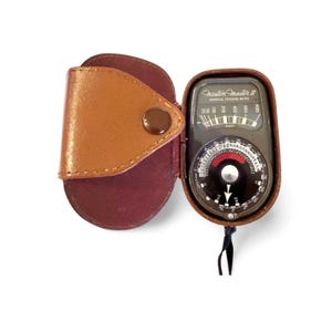 May include: A vintage Weston Master II universal exposure meter in a brown leather case. The meter has a black face with white and red markings, and a needle indicator. The case is open, revealing the meter's dial and needle.