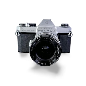 Puede incluir: Una cámara vintage Asahi Pentax Spotmatic F con una lente Soligor wide-auto. El cuerpo de la cámara es plateado y negro, con el nombre de la marca y el número de modelo claramente visibles. La lente tiene una distancia focal de 28 mm y una apertura de f/2.8.