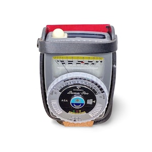 May include: A vintage Gossen Luna-Pro light meter in a leather case. The meter has a gray body with a dial featuring numbers and settings for ASA, time, and f-stops. The top has a red accent.