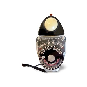 Può includere: Un esposimetro Director Color-Matic vintage. Lo strumento ha un corpo in plastica trasparente con quadranti in bianco e nero e una parte superiore nera con una cupola bianca. Il testo "DIRECTOR Color-Matic" è visibile sul quadrante.