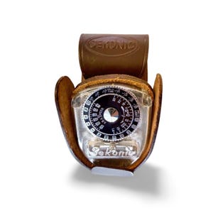 May include: A vintage Sekonic light meter in a brown leather case. The meter has a clear plastic face with a black dial and white markings. The leather case has the Sekonic logo embossed on the top.