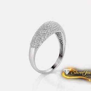 Könnte beinhalten: Ein silberner Ring mit geschwungenem Design, vollständig besetzt mit kleinen, funkelnden weißen Edelsteinen. Der Ring hat einen glatten, polierten Ring und eine abgerundete Oberseite, die mit den Edelsteinen bedeckt ist. Der Ring wird auf weißem Hintergrund präsentiert.