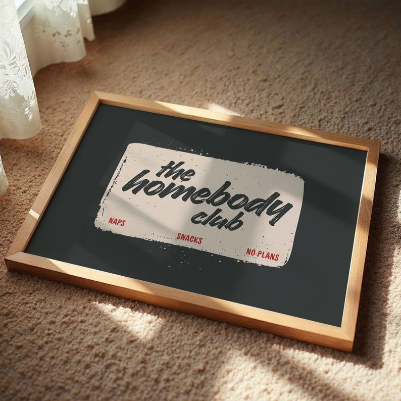 May include: A framed chalkboard with the text "the homebody club" in a vintage style. The words "naps", "snacks", and "no plans" are also visible. The frame is made of wood and the background is a dark gray color.