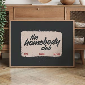 May include: A framed print with the text "the homebody club" in a distressed font. The print has a black background with a beige rectangle in the center. The words "naps", "snacks", and "no plans" are below the text.