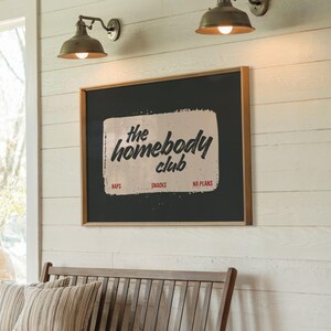 May include: A framed sign with the text "the homebody club" in a distressed font. The sign is black with a cream-colored rectangular shape in the center. Below the text are the words "naps", "snacks", and "no plans".