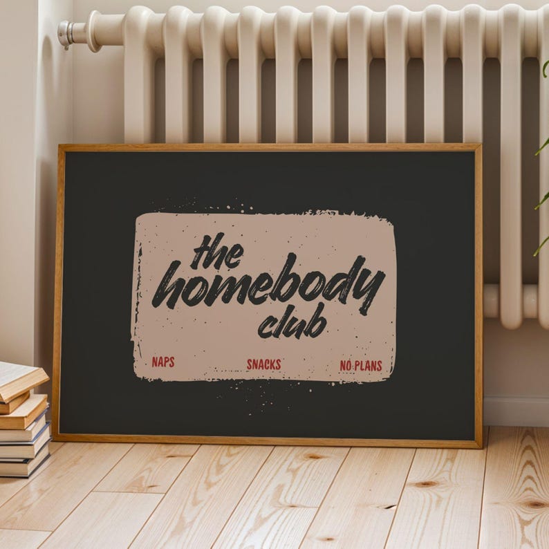 May include: A framed poster with the text "the homebody club" in black script on a tan background. The words "naps", "snacks", and "no plans" are below the text in red. The poster is in a wooden frame.