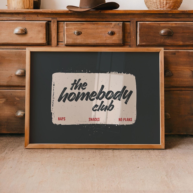 May include: A framed print with the words "the homebody club" in a distressed font. The print has a black background with a beige rectangle containing the text. Below the text are the words "NAPS", "SNACKS", and "NO PLANS". The frame is made of wood.