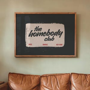 May include: Wooden framed wall art with the text "the homebody club" in a handwritten style. The art piece has a dark background with a light-colored rectangle in the center. Below the text are the words "naps", "snacks", and "no plans".