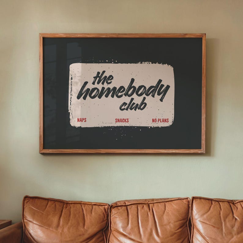 May include: Wooden framed wall art with the text "the homebody club" in a handwritten style. The art piece has a dark background with a light-colored rectangle in the center. Below the text are the words "naps", "snacks", and "no plans".