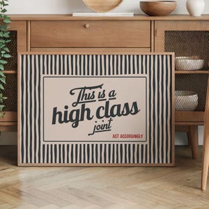 Może przedstawiać: Oprawiony wydruk artystyczny z czarno-białą obwódką w paski. Wydruk zawiera frazę "This is a high class joint" w stylizowanej czcionce, z napisem "Act Accordingly" poniżej. Tło jest w jasnym odcieniu beżu.