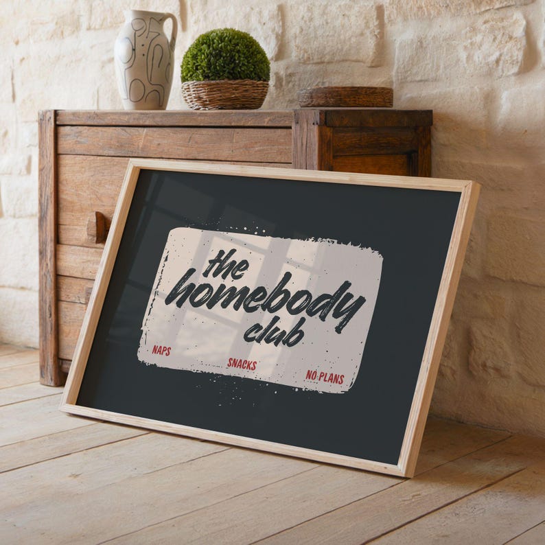 May include: A framed print with the text "the homebody club" in a distressed font. The print is on a dark background with a light-colored rectangle. The words "naps", "snacks", and "no plans" are below the main text. The frame is light wood.