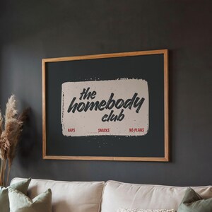 May include: A framed poster with the text "the homebody club" in a handwritten style. The poster has a light beige background with the words "naps", "snacks", and "no plans" below the main text. The frame is made of light brown wood.