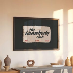May include: A framed print with the words "the homebody club" in a vintage style. The print is black with a cream-colored rectangle containing the text. Below the text are the words "naps", "snacks", and "no plans".