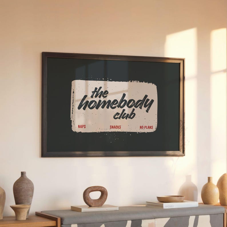 May include: A framed print with the words "the homebody club" in a vintage style. The print is black with a cream-colored rectangle containing the text. Below the text are the words "naps", "snacks", and "no plans".