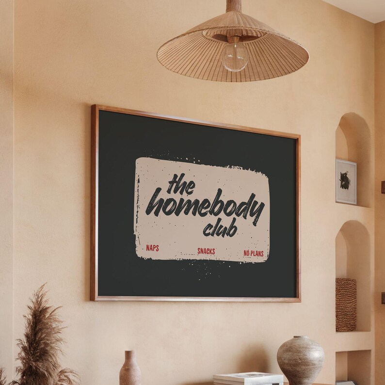 May include: A framed wall art print with the text "the homebody club" in a distressed font. The print includes the words "naps", "snacks", and "no plans". The frame is a dark wood color.