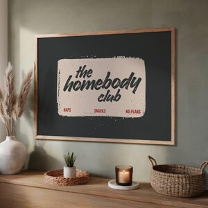May include: A framed poster with the text "the homebody club" in a vintage style. The poster has a dark background with a light-colored rectangle in the center. Below the text are the words "naps", "snacks", and "no plans".