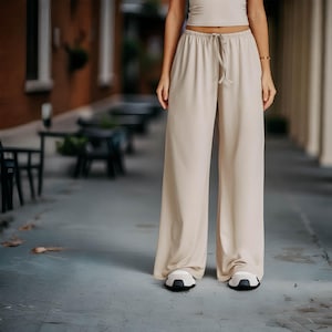 Könnte beinhalten: Beige Hose mit weitem Bein und Kordelzug in der Taille. Die Hose wird mit einem passenden Oberteil kombiniert. Die Person trägt weiß-schwarze Turnschuhe. Das Outfit ist neutral gehalten und scheint aus einem weichen, fließenden Stoff zu sein.