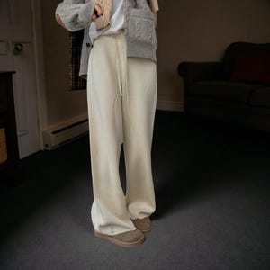 May include: Cream-colored wide-leg sweatpants with a drawstring waist. The pants are paired with brown suede-like shoes. The image is taken indoors, with a neutral background and soft lighting. The pants appear to be made of a soft, comfortable material.