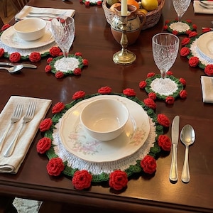 May include: A formal dining table set for a meal. The table is set with white plates, bowls, silverware, and crystal glasses. Each place setting includes a crocheted placemat with red roses and green leaves. A basket of fruit and lit candles complete the scene.