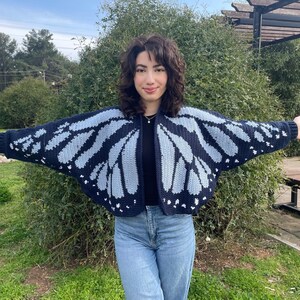 May include: A navy blue and light blue butterfly wing-patterned cardigan. The open-front sweater is made of a knitted material. The cardigan is worn over a black top and paired with blue jeans.