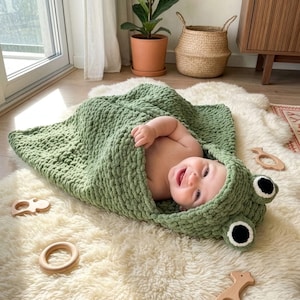 Può includere: Un bambino avvolto in una coperta con cappuccio verde a forma di rana, con occhi neri e bianchi. È sdraiato su un tappeto bianco e soffice, circondato da giocattoli di legno. La coperta è in maglia grossa.
