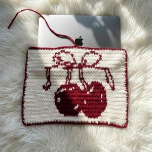 May include: Cream-colored crochet laptop sleeve featuring a pair of red cherries. The sleeve has a red border and a matching red cord handle. The design is set against a fluffy white background.