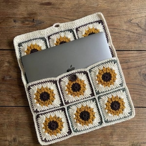 Peut inclure: Housse d'ordinateur portable crochetée avec un motif de tournesol. La housse est faite de fil crème, jaune, marron et vert, avec une fermeture à bouton. Un ordinateur portable argenté est à l'intérieur.