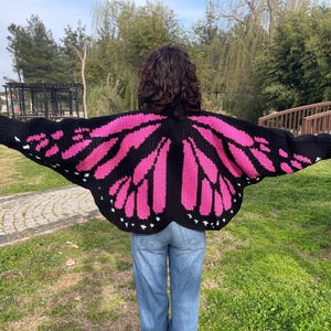 May include: A black and hot pink crocheted cardigan featuring a butterfly wing pattern on the back and sleeves. The garment has long sleeves, a wide silhouette, and small white accents along the bottom edge of the wing design.