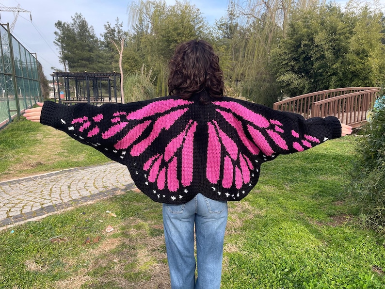 May include: A black and hot pink crocheted cardigan featuring a butterfly wing pattern on the back and sleeves. The garment has long sleeves, a wide silhouette, and small white accents along the bottom edge of the wing design.