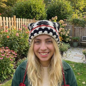 May include: A handmade crochet hat with a black and white pattern, red trim, and ear flaps. The hat has red heart-shaped accents hanging from the sides. The person is wearing a green corduroy jacket and a cream-colored sweater.