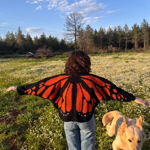 May include: A black and orange butterfly wing-shaped crochet sweater. The sweater features a detailed design with orange wings outlined in black. The person is wearing blue jeans and standing in a field of white flowers with a golden retriever dog.
