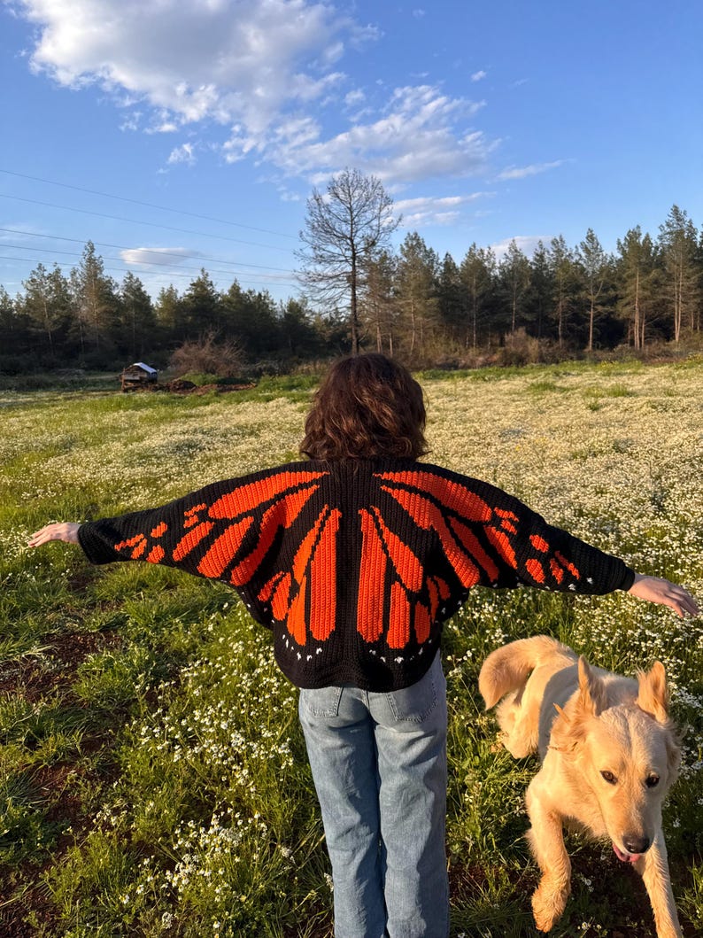 May include: A black and orange butterfly wing-shaped crochet sweater. The sweater features a detailed design with orange wings outlined in black. The person is wearing blue jeans and standing in a field of white flowers with a golden retriever dog.
