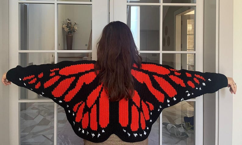 May include: A black crochet cardigan featuring a vibrant red butterfly wing pattern across the back and sleeves. The design includes distinct red segments outlined in black, with small white accents along the edges of the wings.
