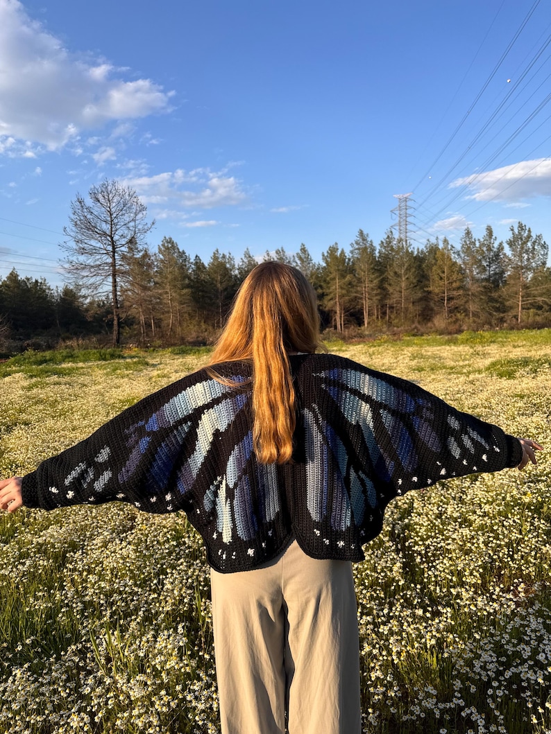 May include: A black crochet butterfly wing cardigan with blue and white accents. The open-front sweater features a detailed wing design, perfect for layering. The cardigan is worn outdoors in a field of white flowers.